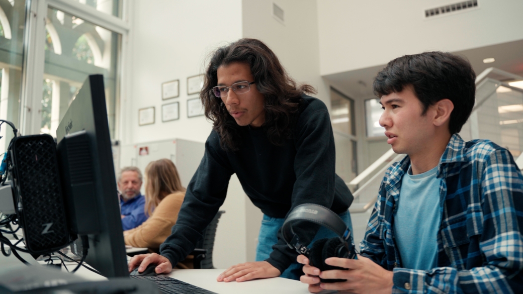 A student bends over a computer, helping another student with his work.