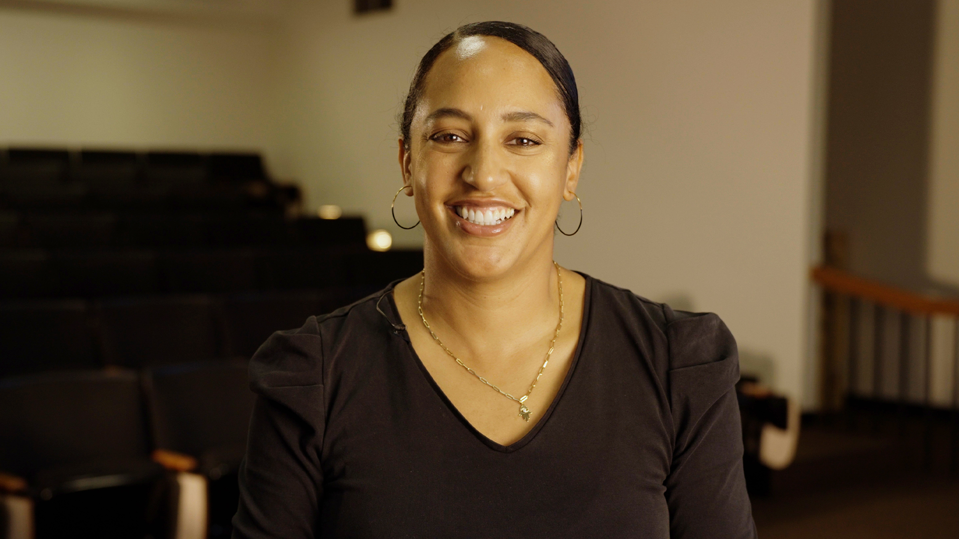Melanie Cherry smiles at the camera wearing a black shirt in front of an empty auditorium/classroom. 