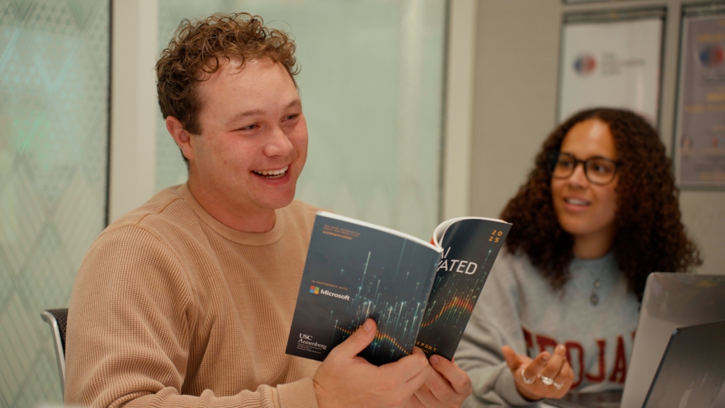 A student smiles while holding open a research report and another student looks on. 