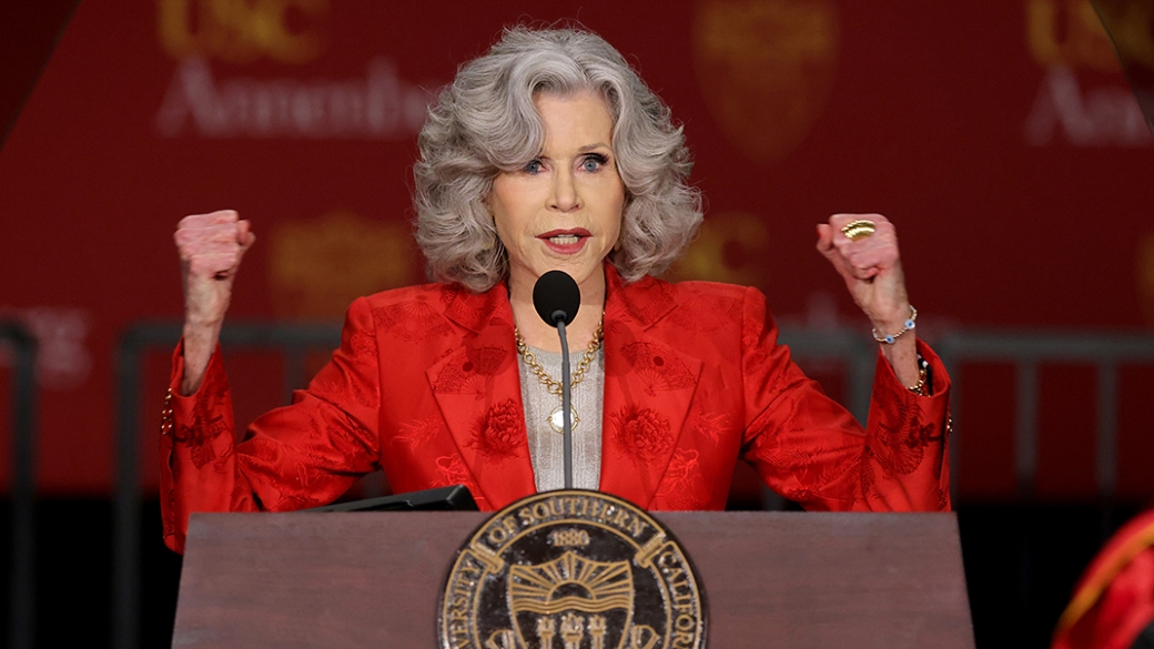 Wearing a red suit, Jane Fonda raises two arms, imitating a muscle flex, in front of a podium.