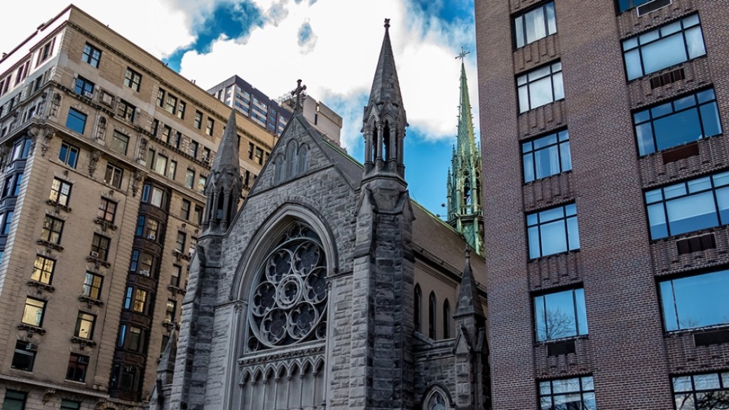 A 3/4ths view of a cathedral building sandwiched between two larger metropolitan buildings on a sunny day