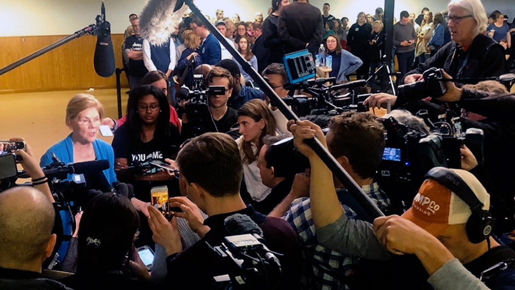 A crowd of reporters surround Elizabeth Warren, who is speaking. 