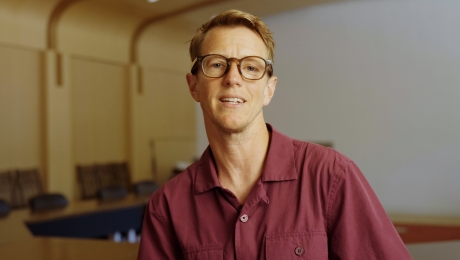 Rook Campbell wears a red shirt and glasses and speaks to the camera in front of an empty classroom.