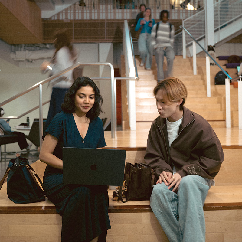 Two students sit together on indoor steps, looking at a laptop, while other students walk down the staircase behind them.