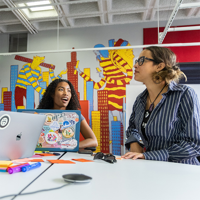 Two women work together at a desk, with a laptop between them
