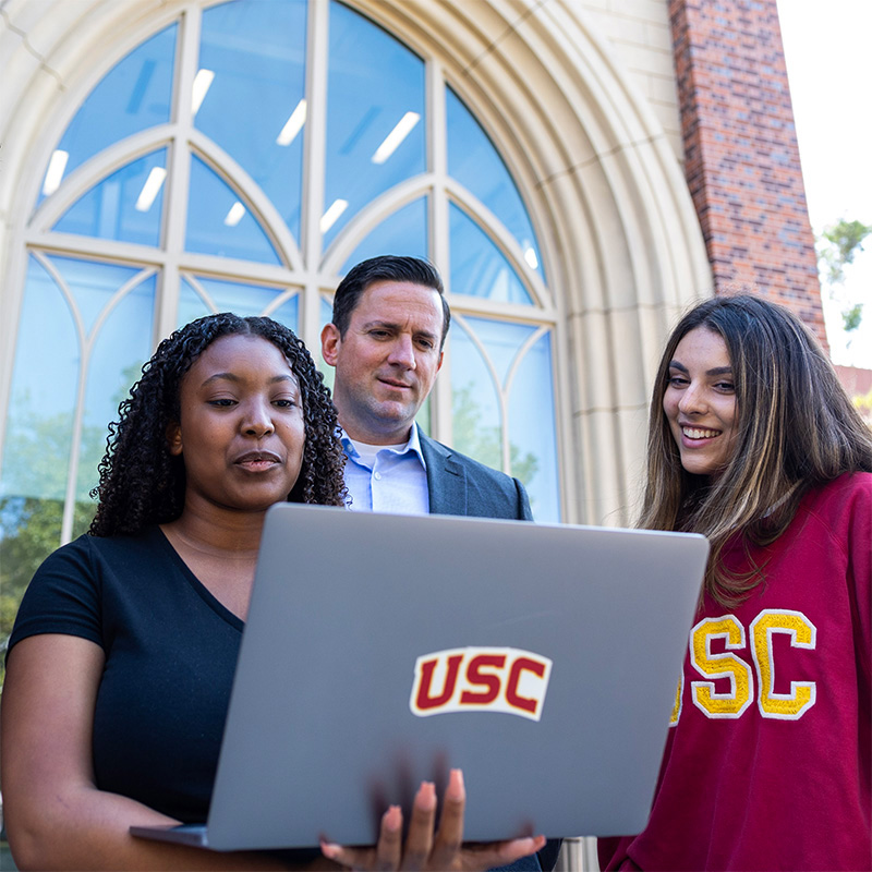 Three people outside of ANN work together on a laptop with a USC sticker on it