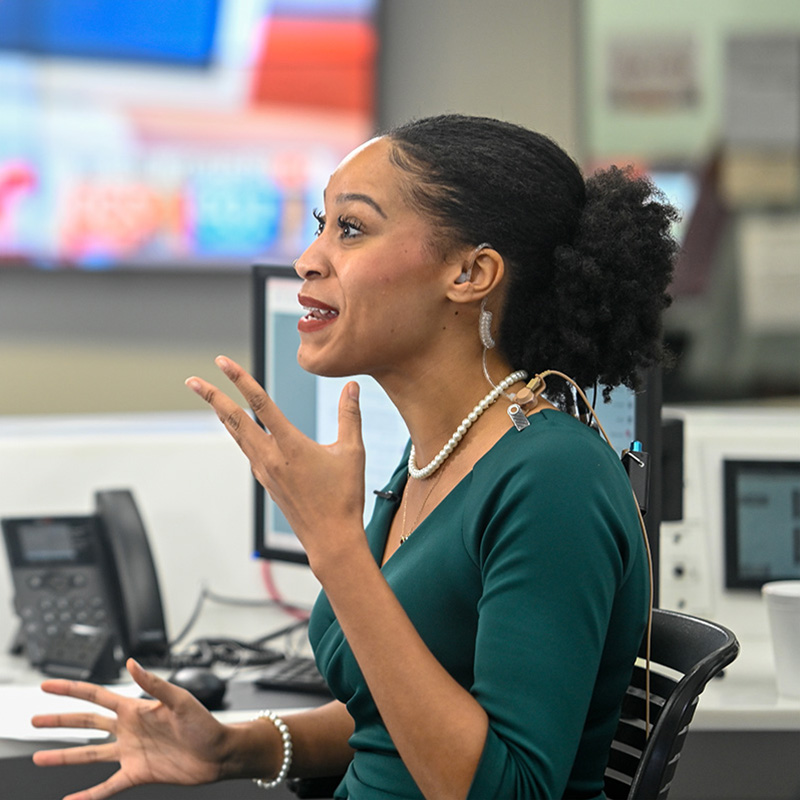 A woman sitting at a desk speaks to someone offscreen