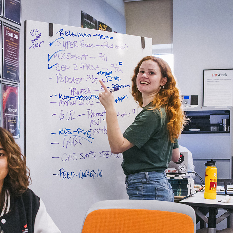 A woman stands writing out ideas on a whiteboard, smiling at the camera