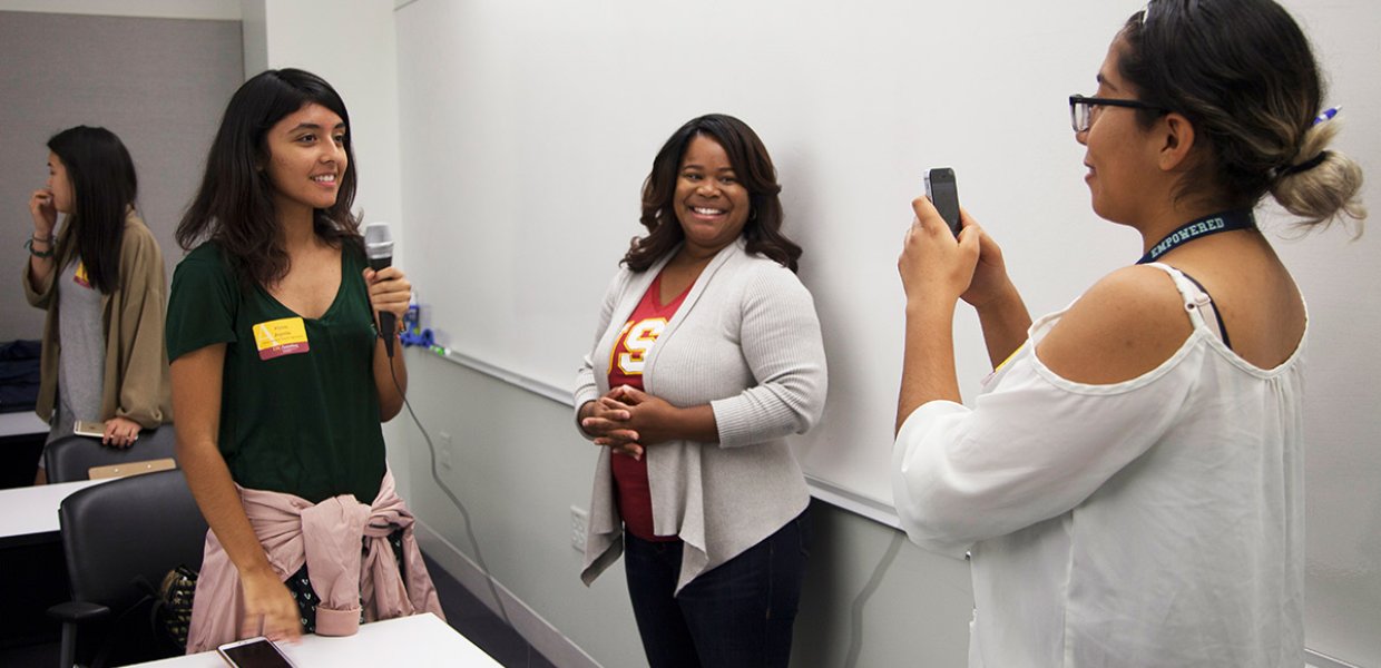 Hispanic female college student holding microphone stands on left of Black female professor while another Latina students films on phone  
