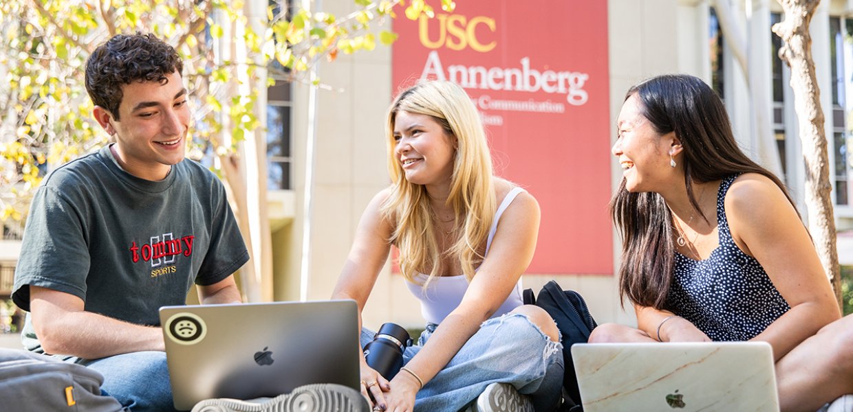 A group of 3 USC Annenberg students study outside with laptops in front of the USC Annenberg building.