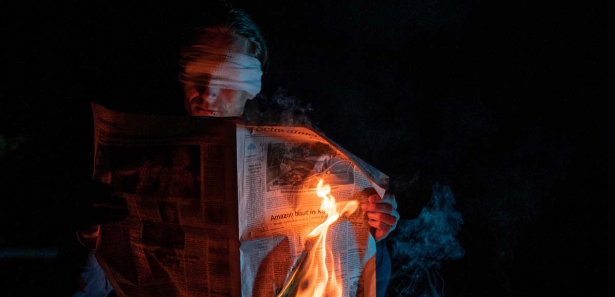 Image of a person wearing a blindfold holding a newspaper that is on fire