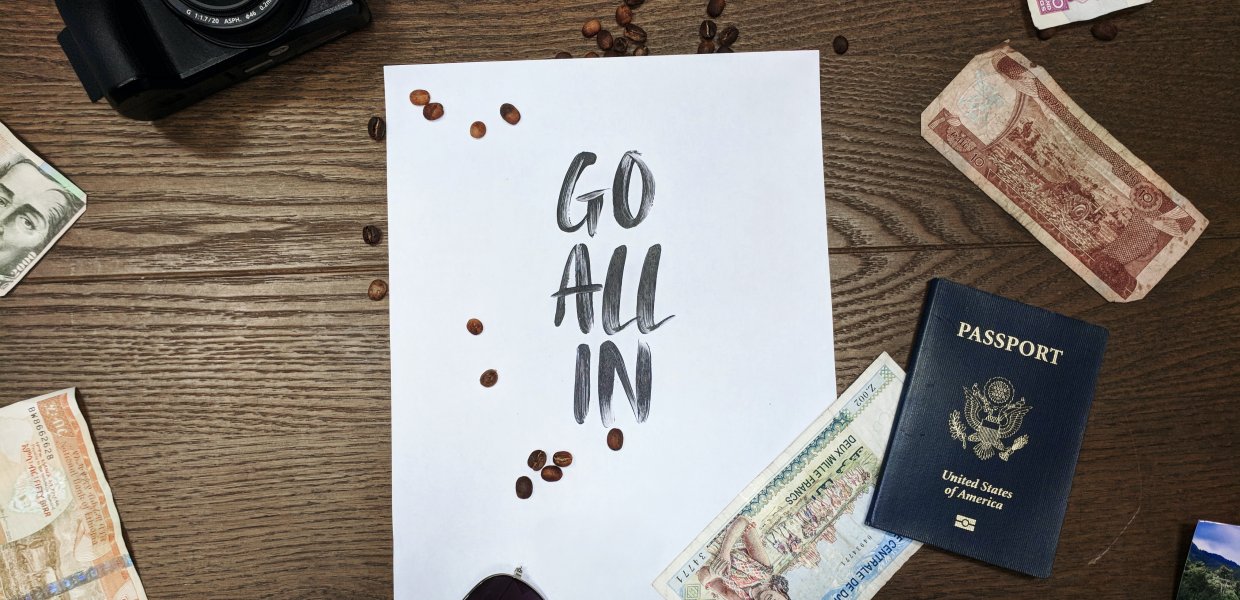 Photo of a piece of paper that reads "go all in" surrounded by sunglasses, a camera, a passport, and different money notes