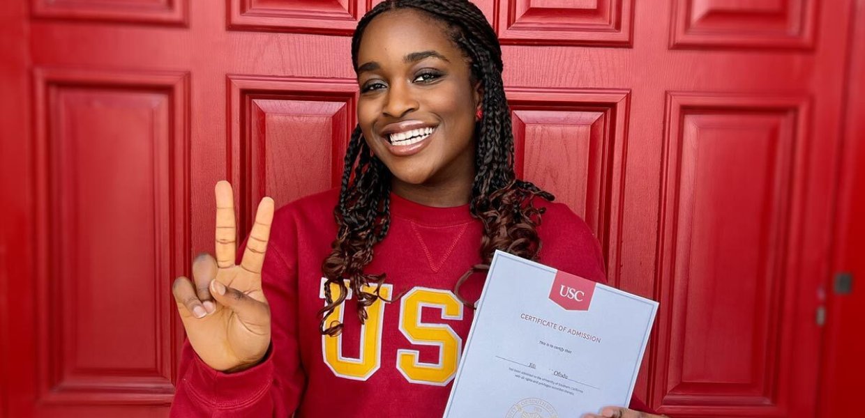 black female college student wearing red sweater holds up victory hand sign while showing acceptance letter in front of red door