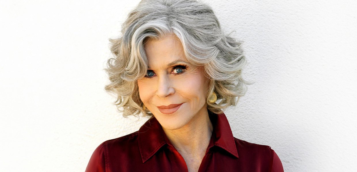 Activist and actor Jane Fonda smiles at camera, wearing a red collared shirt against a white background. 