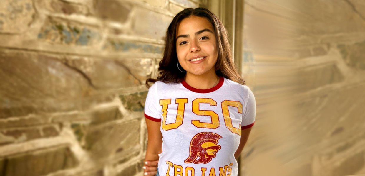 Incoming USC Annenberg master's student, Jenisty Colón, celebrates getting into USC by posing near a building with a USC t-shirt on.