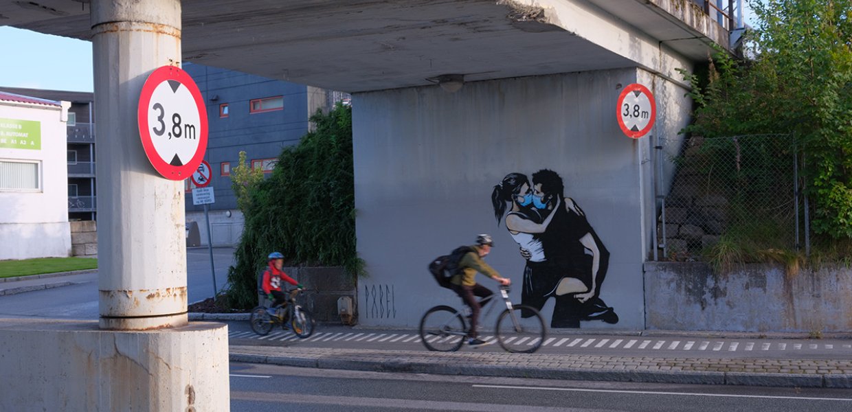 Two people on bicycles under a highway that has an illustration painted on of two people kissing with medical masks on