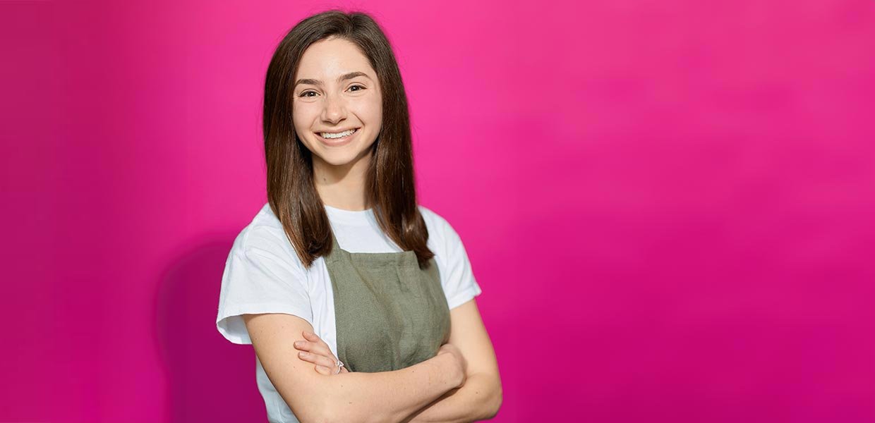 woman in early 20s wearing cooking apron poses in front of pink background