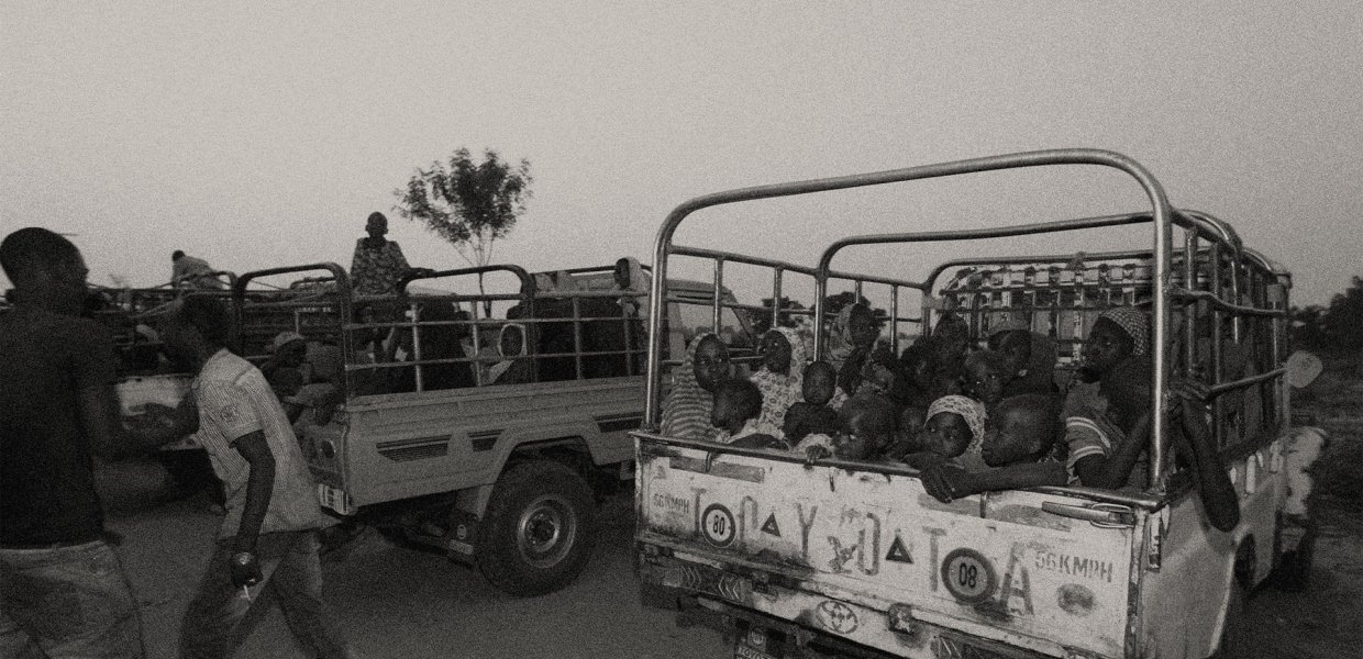 black and white photo of Nigerian women and children scared in back of truck as men walk around guarding