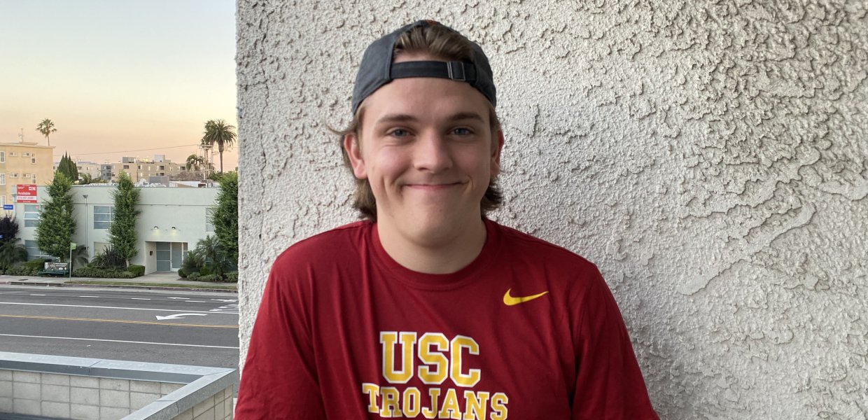 Jack Brockman, a new freshman at USC stands against the wall with a red and gold-lettered USC Trojans shirt and black backwards cap.