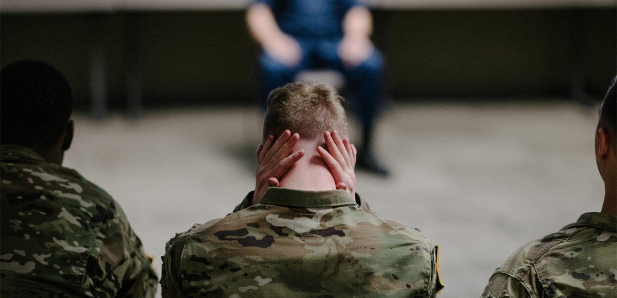 Photo of a military member with their hands on the back of their neck, sitting next to other members
