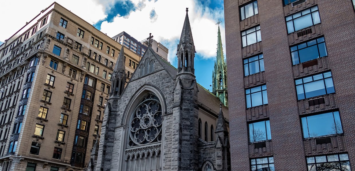 A 3/4ths view of a cathedral building sandwiched between two larger metropolitan buildings on a sunny day