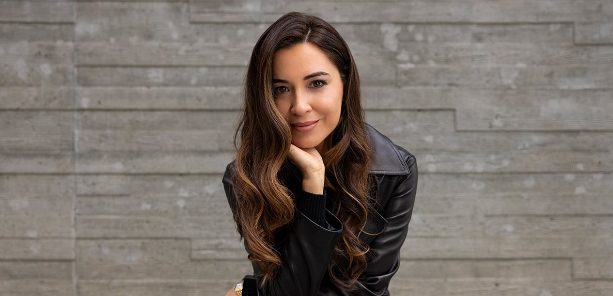 Woman wearing all black sits on a bench in front of a stone wall. She is leaning forward with her elbow on one knee, smiling slightly. 