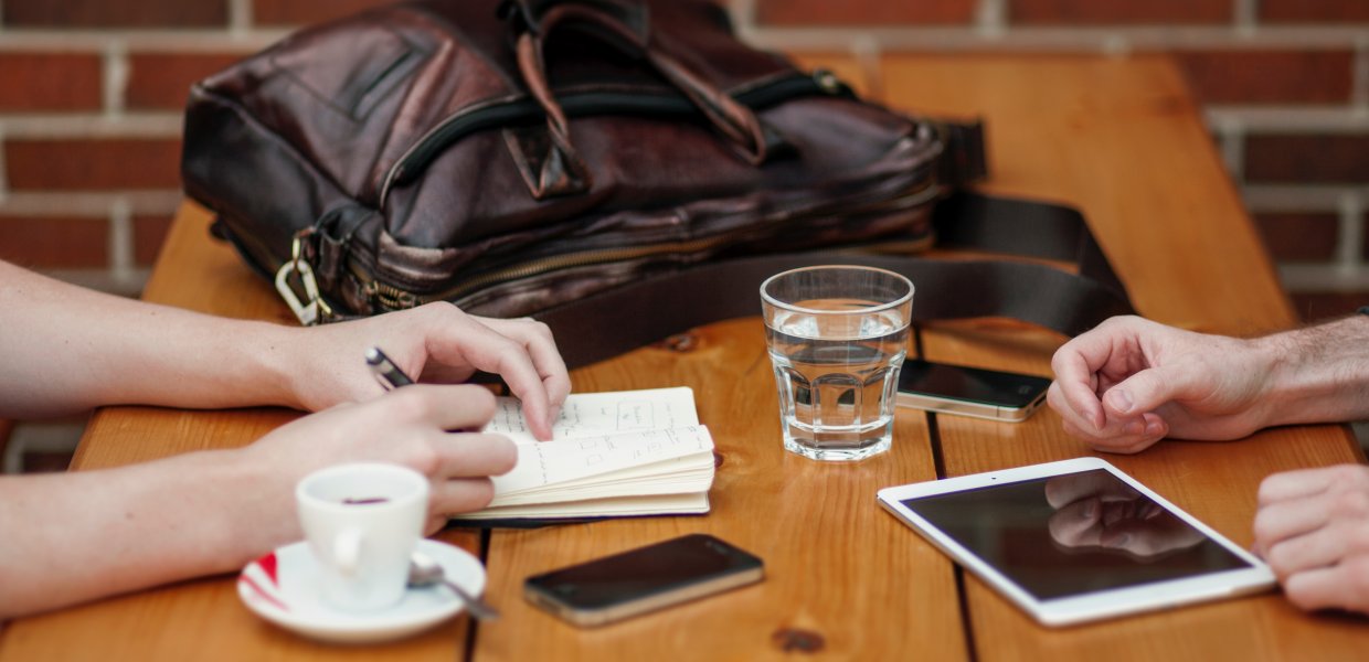 Photo of two people sitting around a table with notepads, IPads, and a coffee cup and water cup