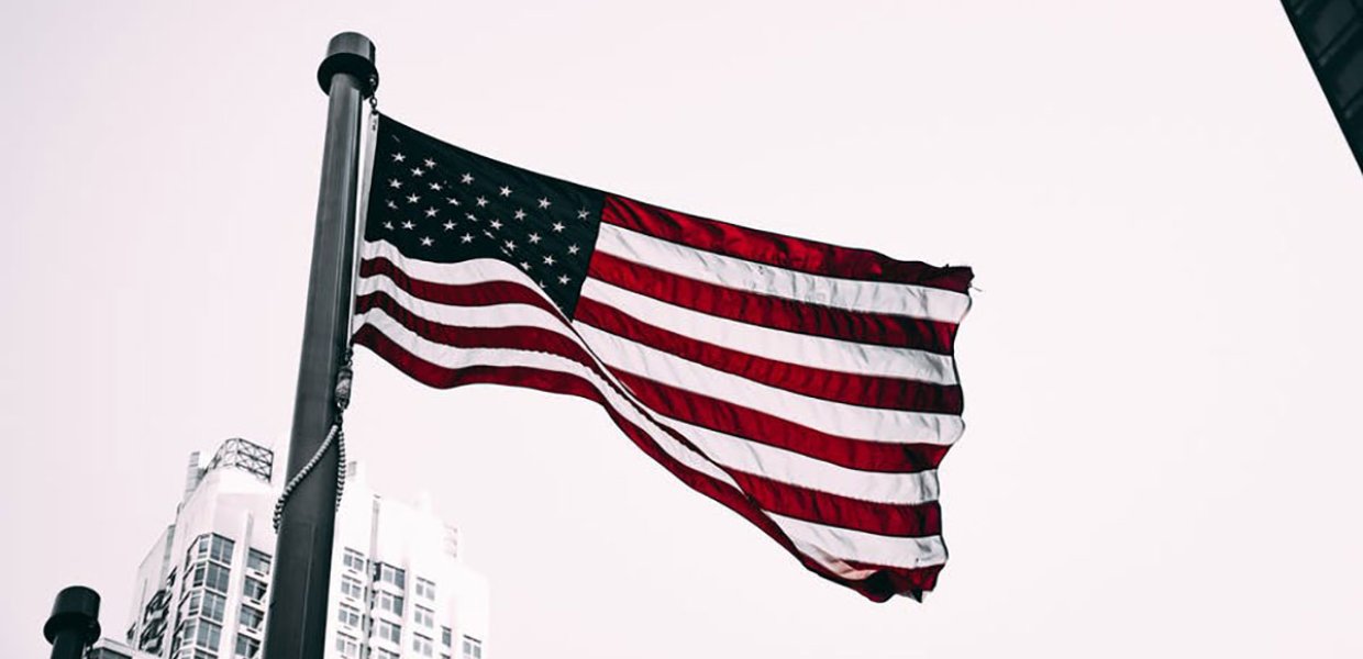 Photo of an American flag in front of a white building with a cloudy sky as a backdrop