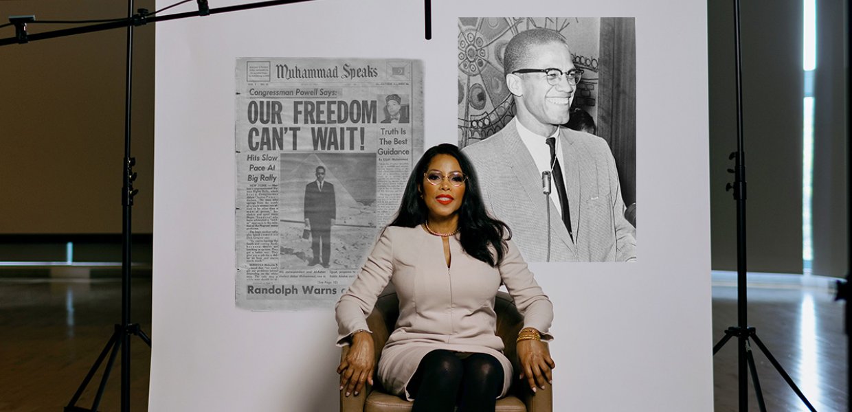 Woman in chair in front of newspaper and photo of Malcolm X