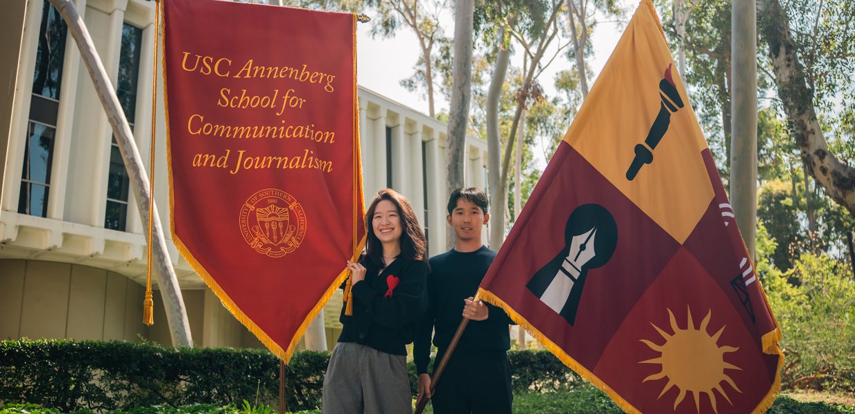 Two individuals stand in front of the Annenberg School for Communication Building holding flags. On the left, a woman holds a cardinal flag bearing the name of the school, and on the right, a man holds a flag bearing symbols that represent the school.