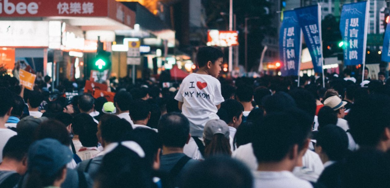 Photo of a crowd at the Hong Kong Protest and a kid on someone's shoulders wearing a shirt that said "I <3 my mom"