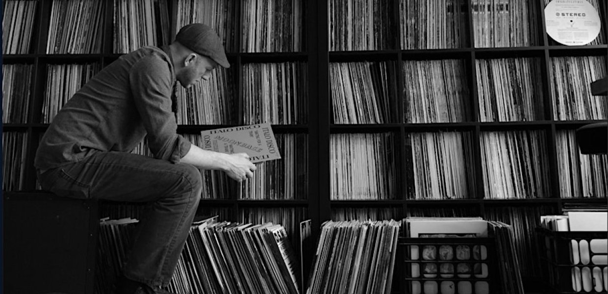 Black and white photo of a man sitting in front of a shelf of records, observing one of them.