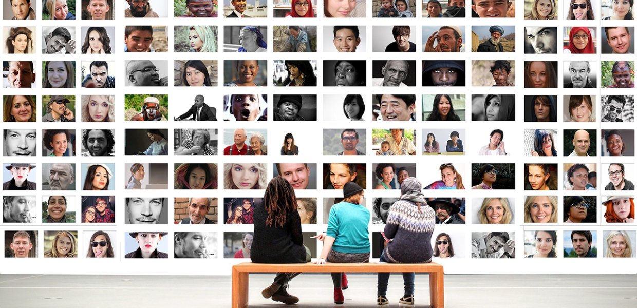 Image of three people sitting on a bench in front of a wall featuring the faces of many different people