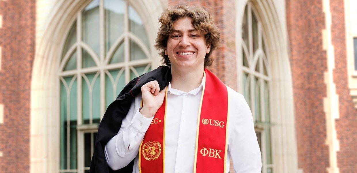 male college graduate smiles for camera wearing red and gold stole and holding graduation robe over one shoulder