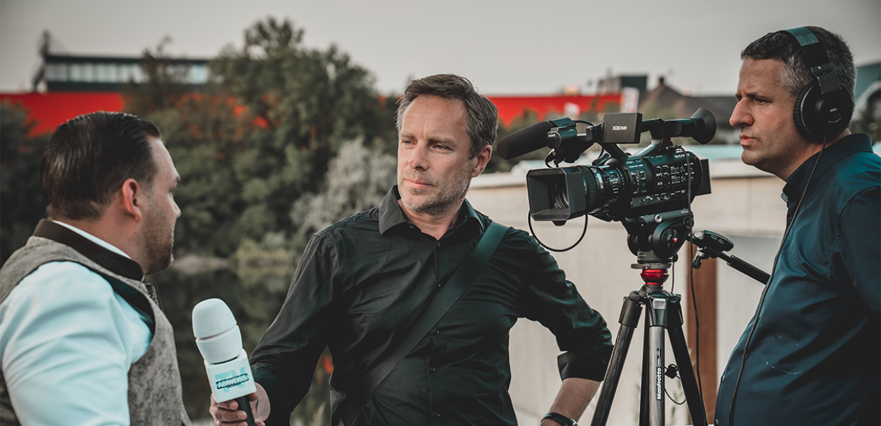 Photo of a man holding a microphone while talking to another man and being filmed