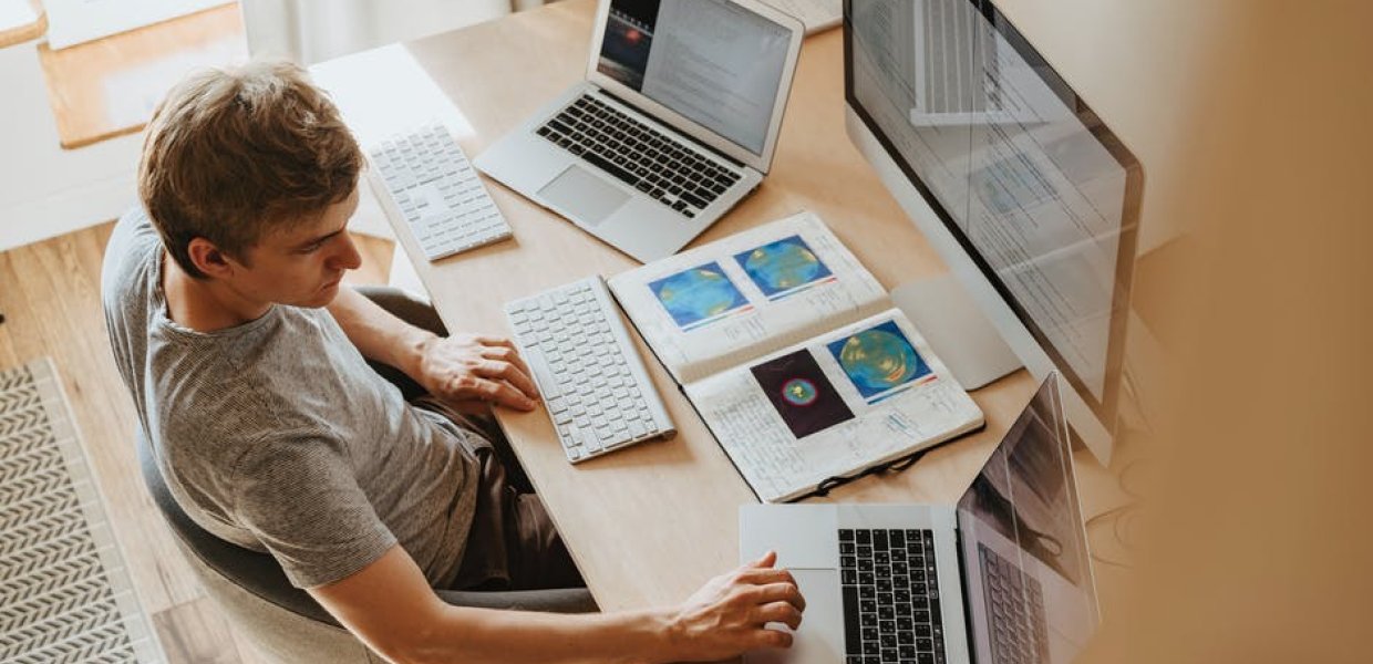 Photo of a person with two laptop computers on either side of them, an open book in front of them, and a desktop computer in front of them