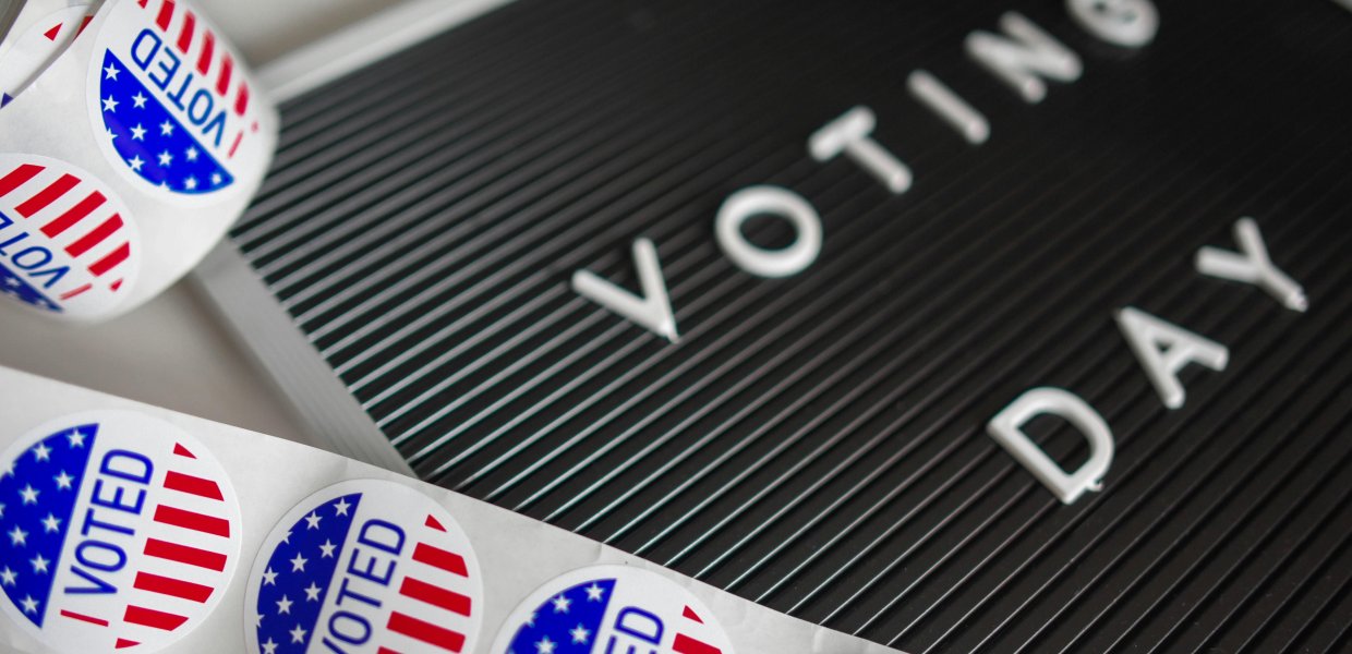 A close-up image of a blackboard with the words “VOTING DAY” spelled out in white capital letters. In the foreground, there is a roll of circular stickers with an American flag design and the text “I VOTED” prominently displayed.