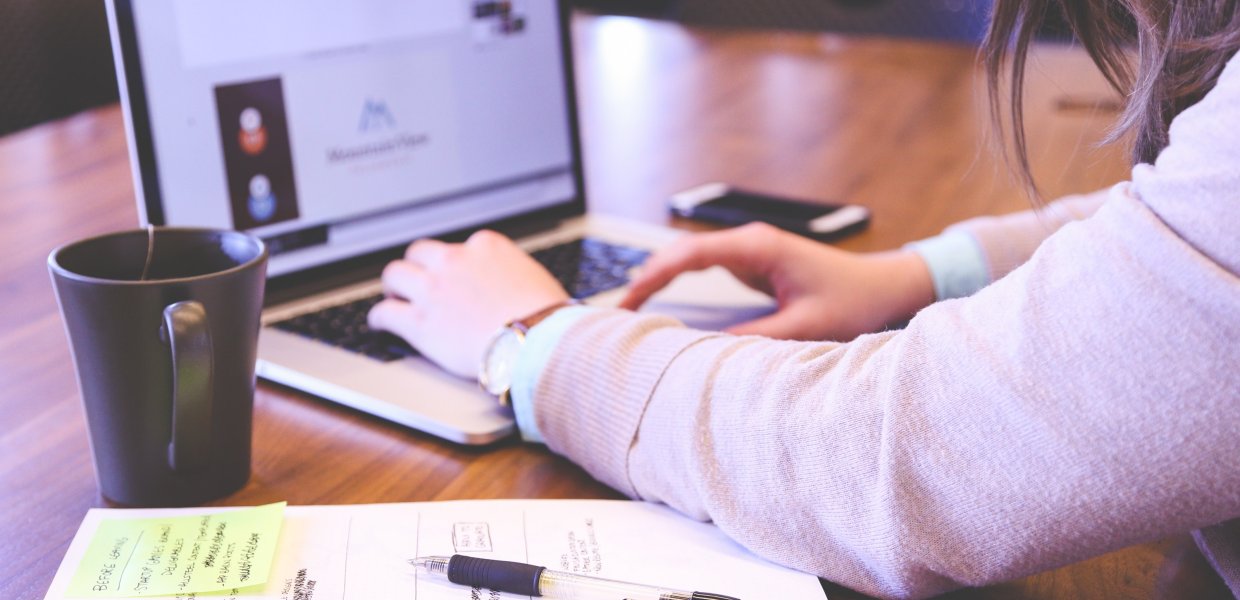 Photo of a person using a computer next to some handwritten notes and a mug