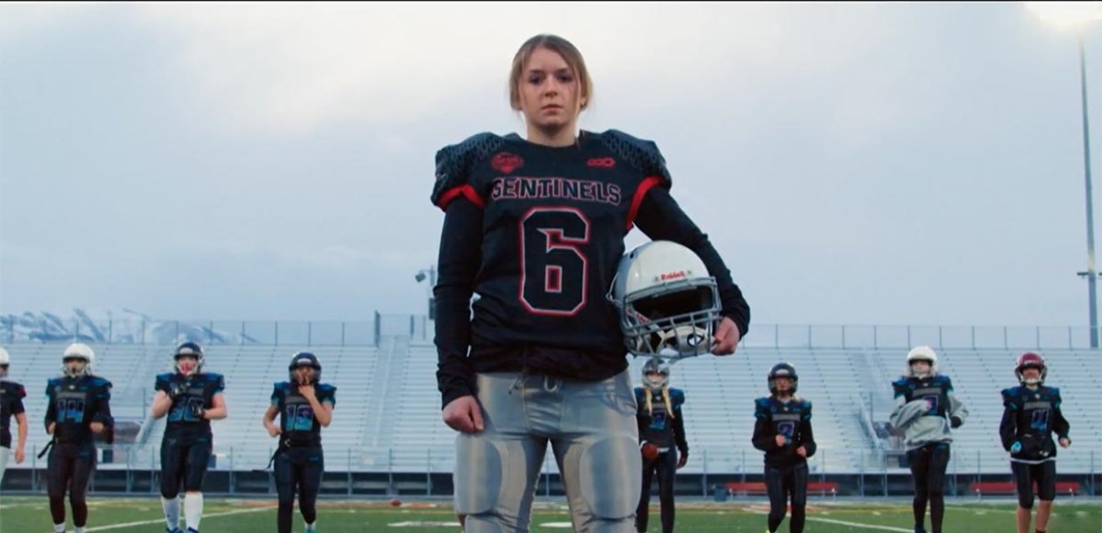 Young girl in football uniform standing in a field, looking at the camera as other girls train behind her.