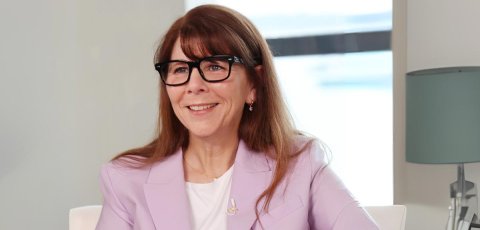 Stacy Smith smiling in front of a window wearing a light pink blazer. 