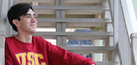 Young man sitting on white steps wearing a cardinal USC sweater.