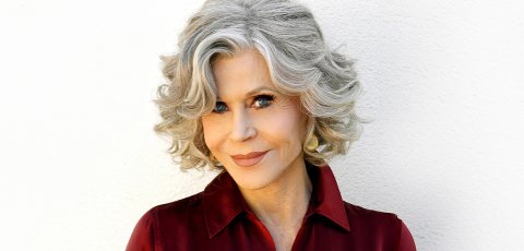 Activist and actor Jane Fonda smiles at camera, wearing a red collared shirt against a white background. 