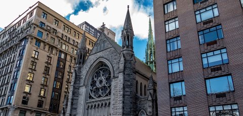A 3/4ths view of a cathedral building sandwiched between two larger metropolitan buildings on a sunny day