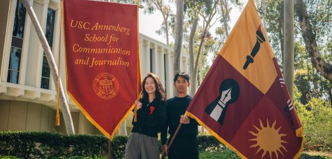 Two individuals stand in front of the Annenberg School for Communication Building holding flags. On the left, a woman holds a cardinal flag bearing the name of the school, and on the right, a man holds a flag bearing symbols that represent the school.