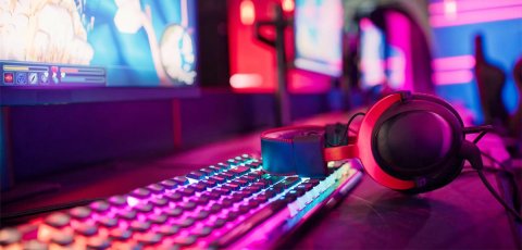 A set of headphones rests on a keyboard, which is in front of a computer screen that displays a video game. The lights are colorful and evoke cyber space. 