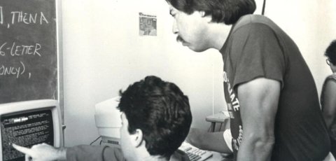 A black and white photo of two men looking at a computer screen. One of the men is pointing to code on the screen. 