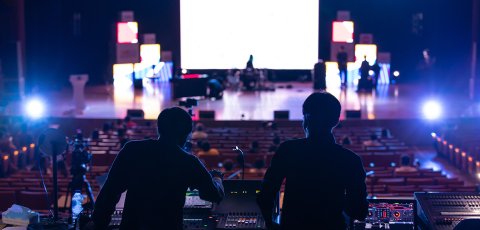 Two men sit at a control panel as a live performance is set up on stage in front of them with a huge white screen.