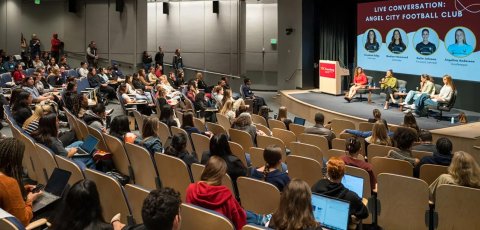 A full auditorium watches a panel of four women soccer players.