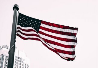 Photo of an American flag in front of a white building with a cloudy sky as a backdrop