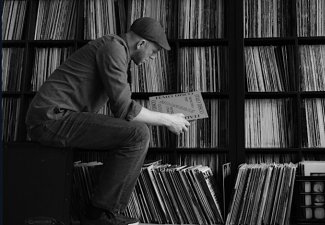 Black and white photo of a man sitting in front of a shelf of records, observing one of them.